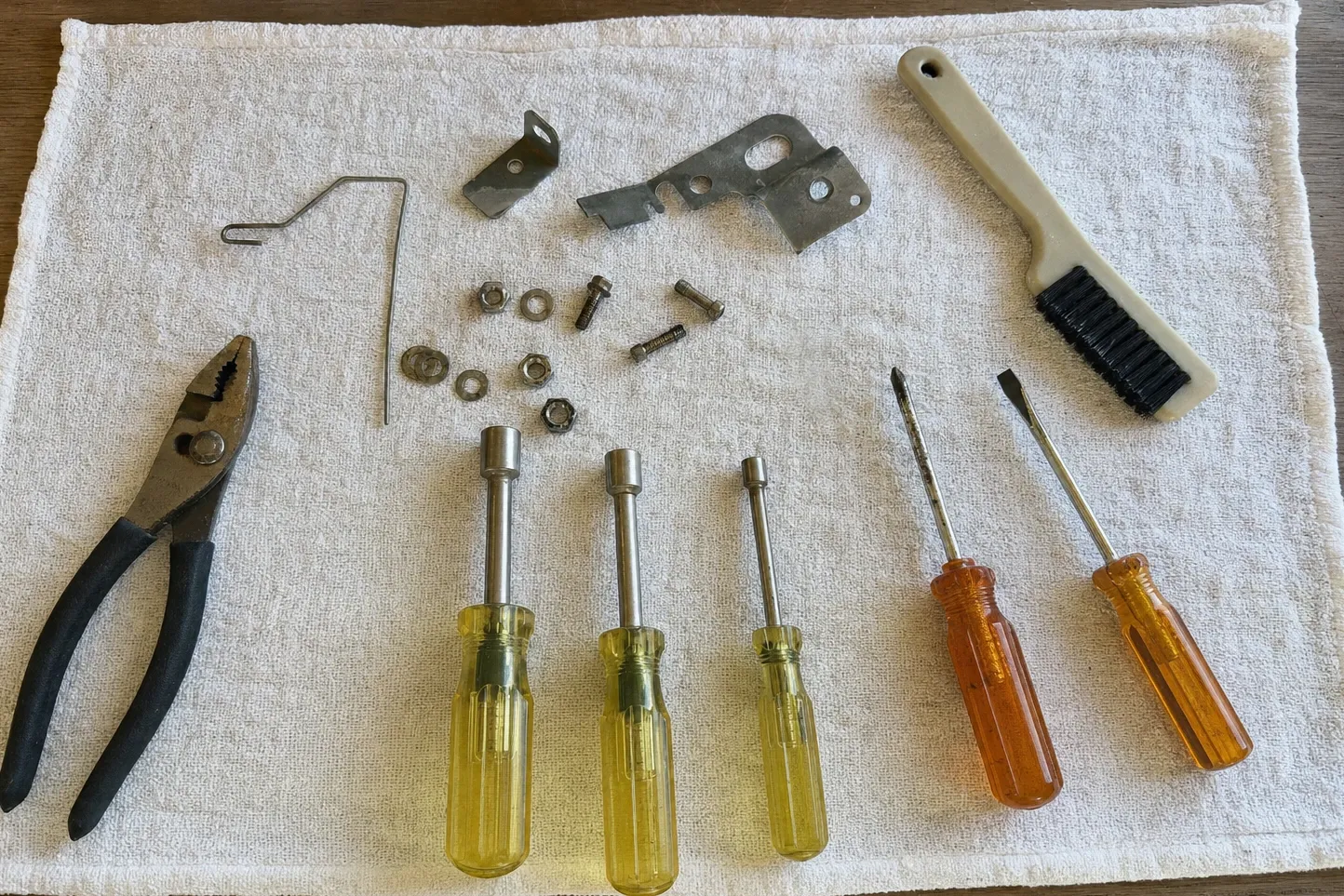 Slip-joint pliers, a bent wire retaining clip, two sheet-metal brackets, three nut drivers and a wire brush on a white towel