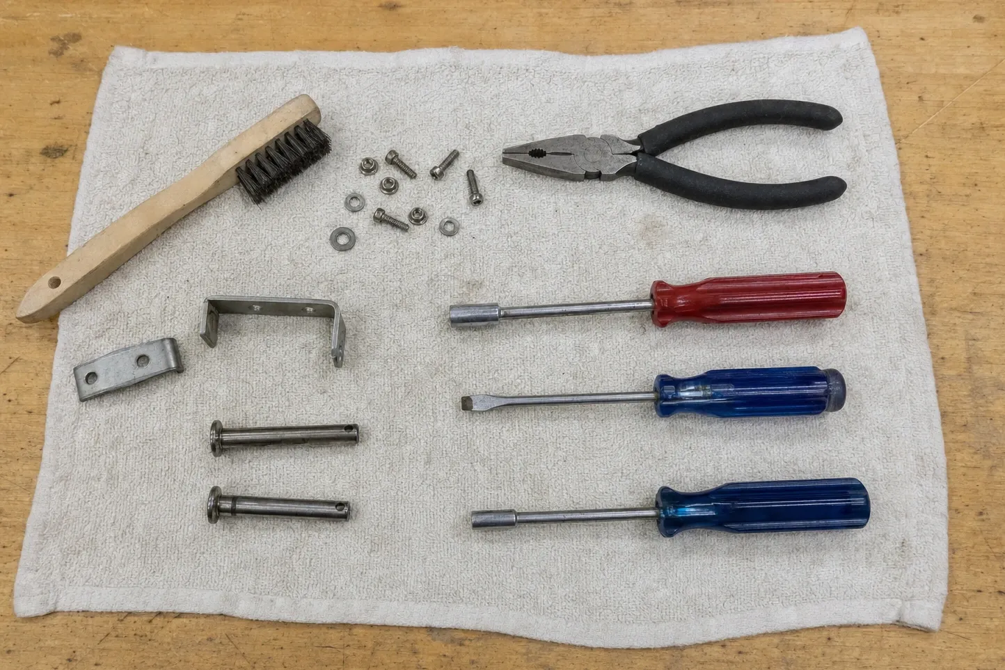Two clevis pins, a flat strap plate and a U-shaped bracket laid out with nut drivers, pliers and a wooden wire brush